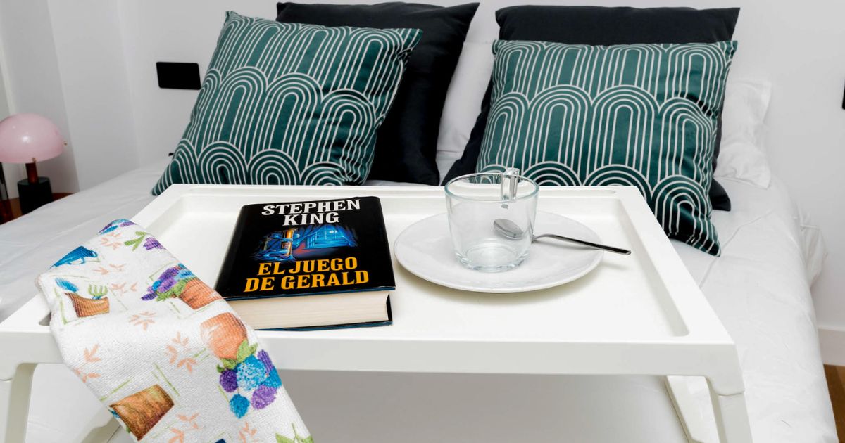 Bandeja blanca sobre la cama con un libro una taza de cristal y un plato con cuchara