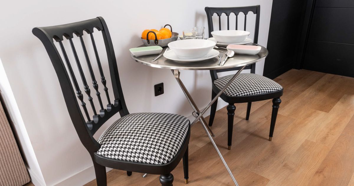 Mesa redonda con dos sillas negras, vajilla blanca y frutero sobre suelo de madera reformado.