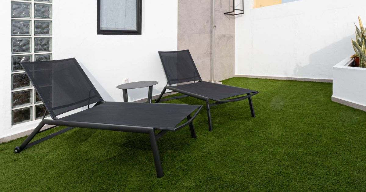 Terraza exterior con césped artificial dos tumbonas negras y mesa en un espacio reformado.